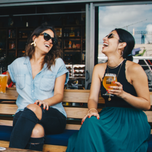 two women sitting on bench holding beer glasses and laughing