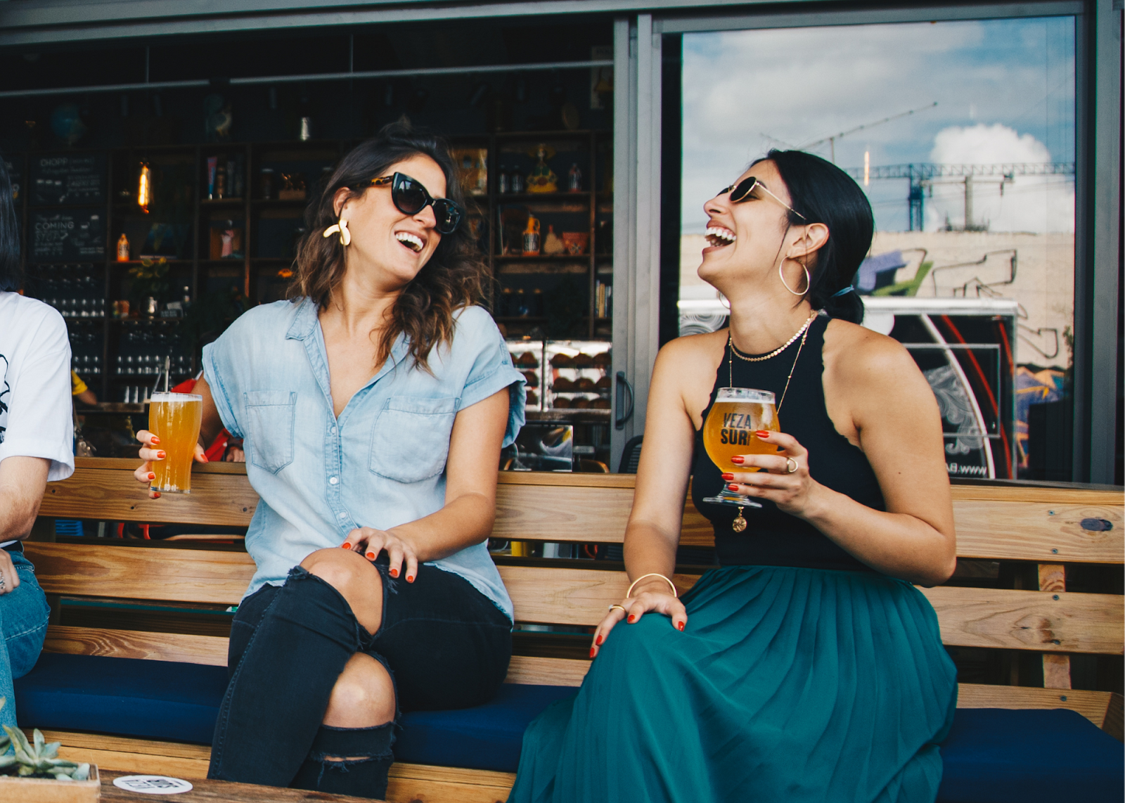 two women sitting on bench holding beer glasses and laughing